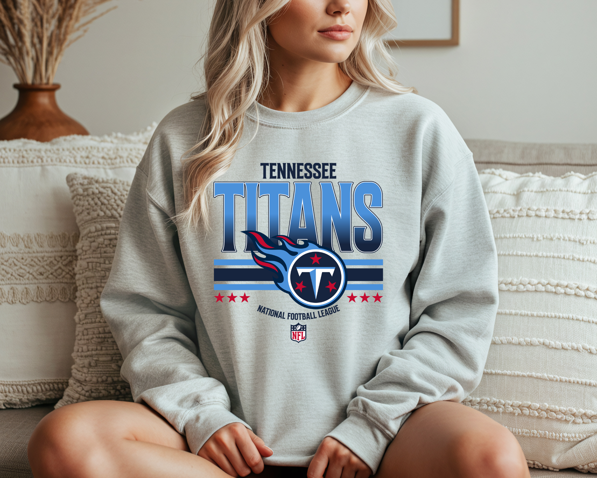 A woman wearing a light gray Tennessee Titans-inspired crewneck sweatshirt with bold blue and red team-style graphics, sitting comfortably on a couch.