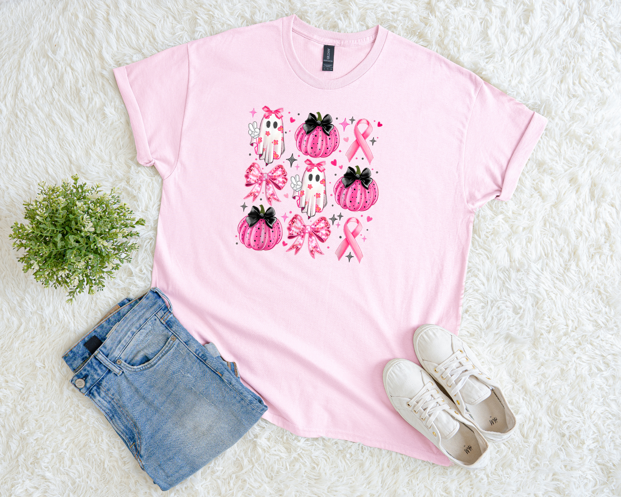 A light pink t-shirt featuring a grid of pink pumpkins, bows, ghosts, and breast cancer ribbons, styled flat with jeans, white sneakers, and a small green plant on a soft white background.