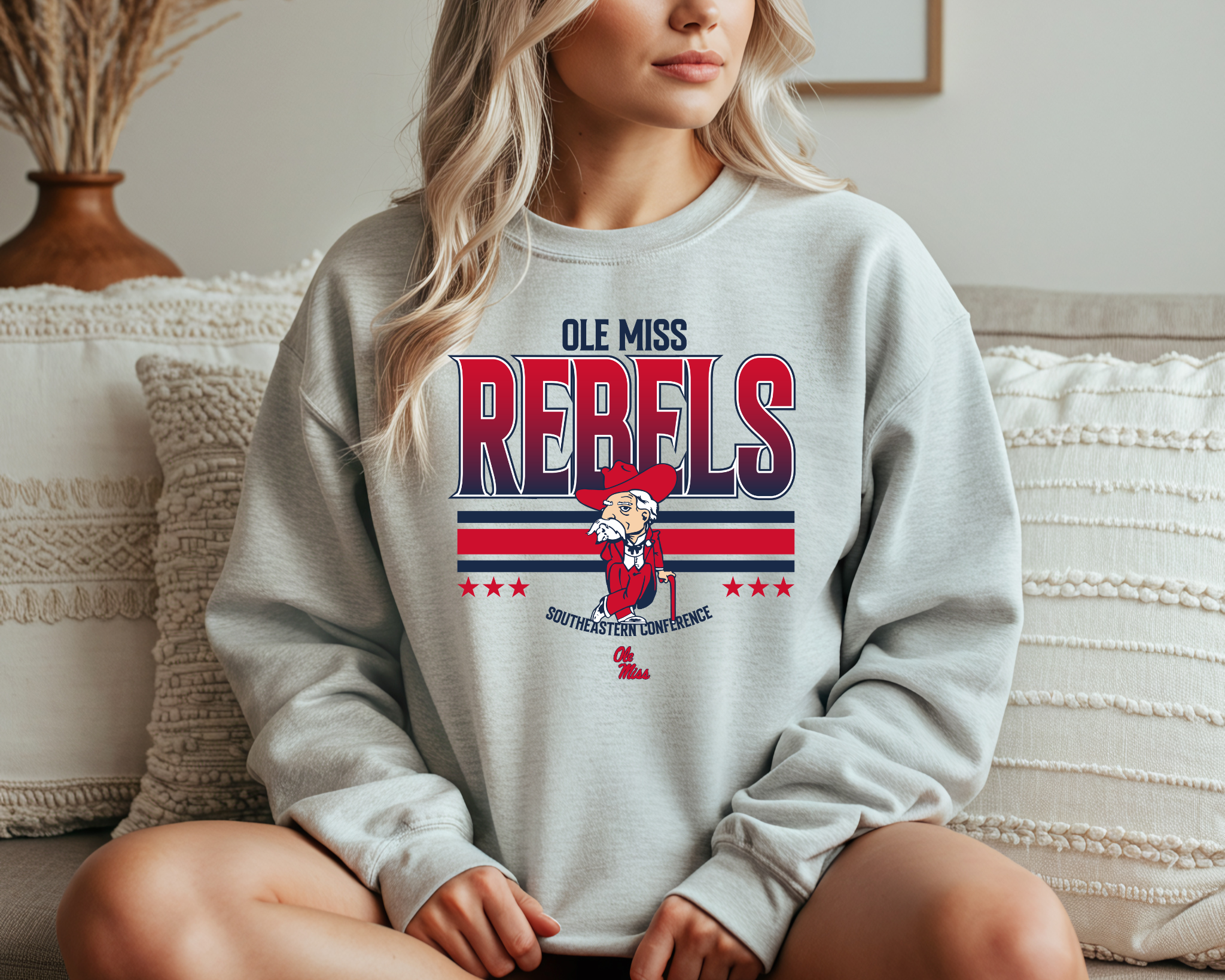 A woman wearing a light gray Ole Miss Rebels-inspired crewneck sweatshirt with bold red and navy graphics and a vintage-style mascot design, sitting comfortably on a couch.