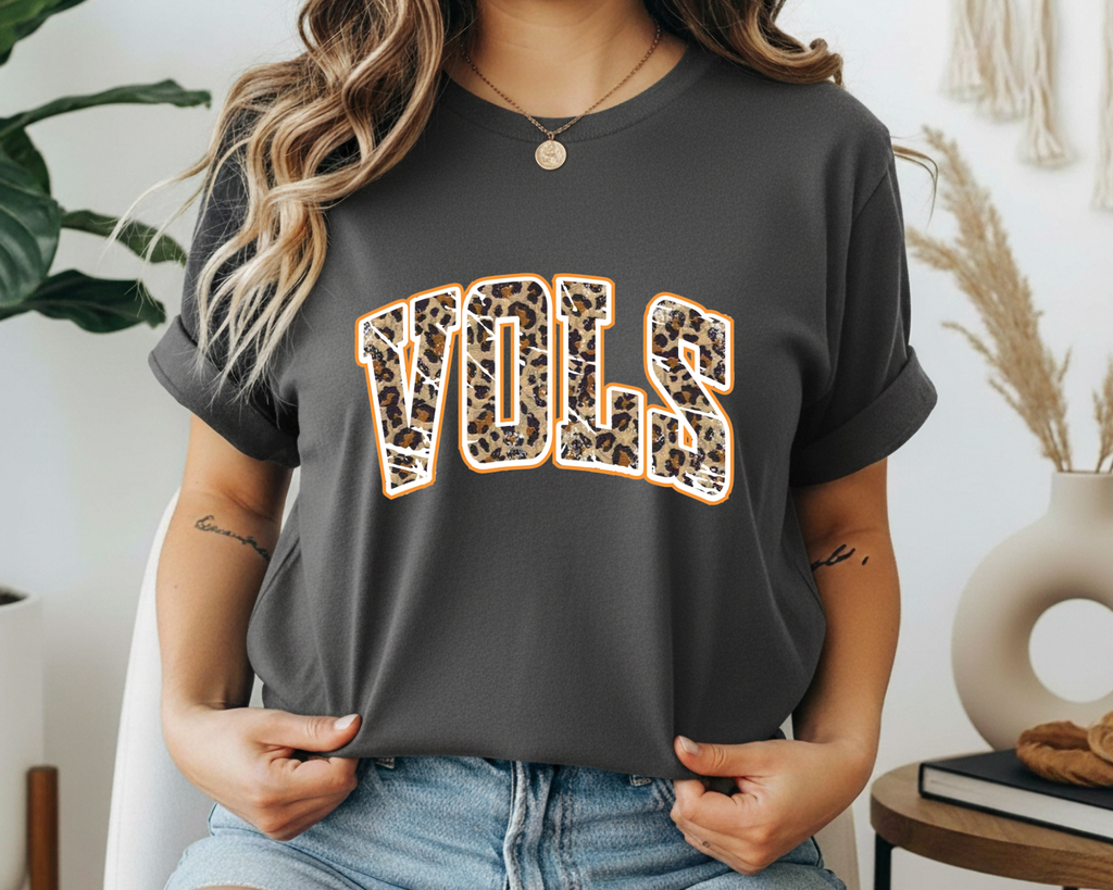 Person wearing a dark gray t-shirt with 'VOLS' printed in leopard print, sitting indoors.