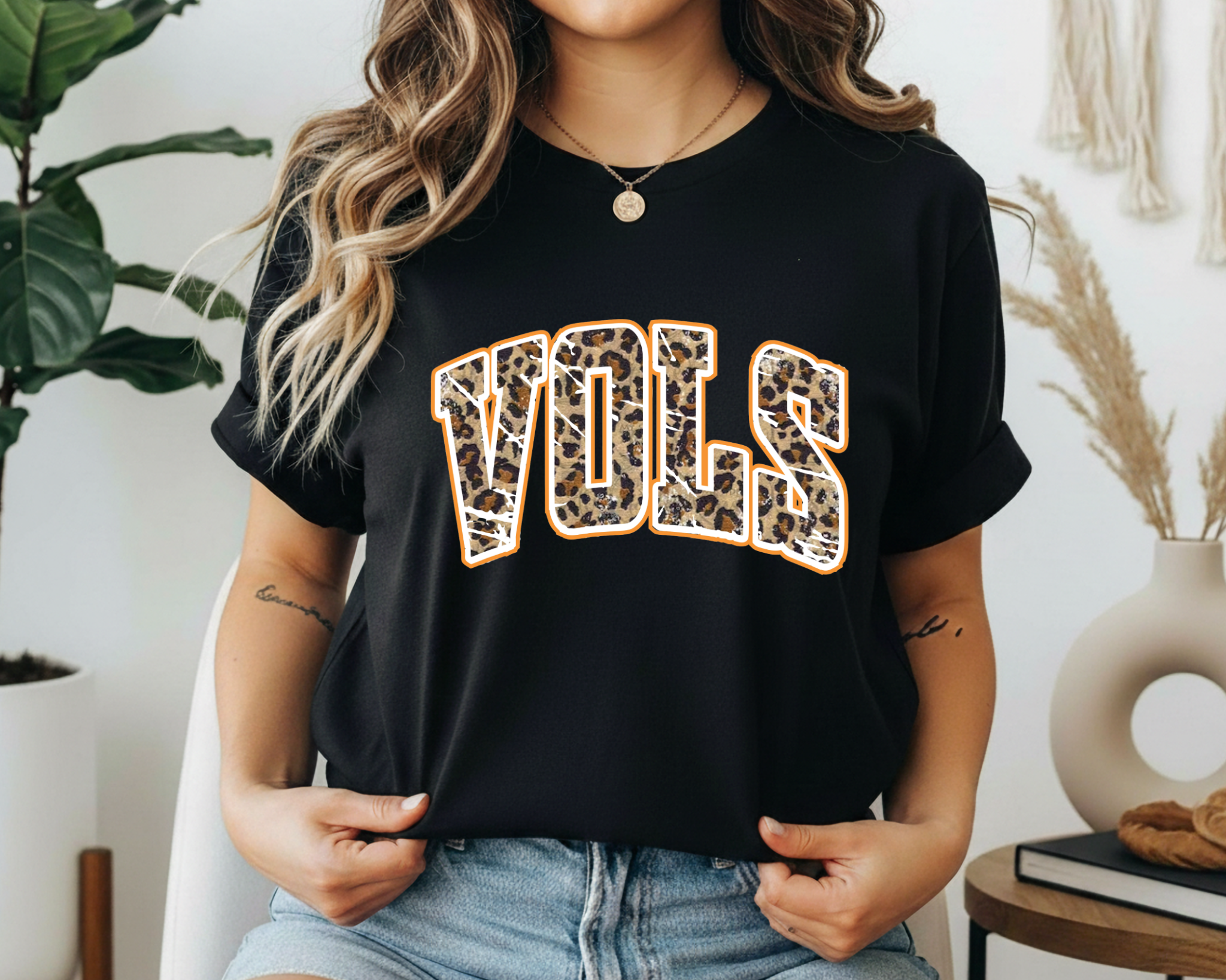 Person wearing a black t-shirt with 'VOLS' printed in leopard print, standing in a room with plants and decor.