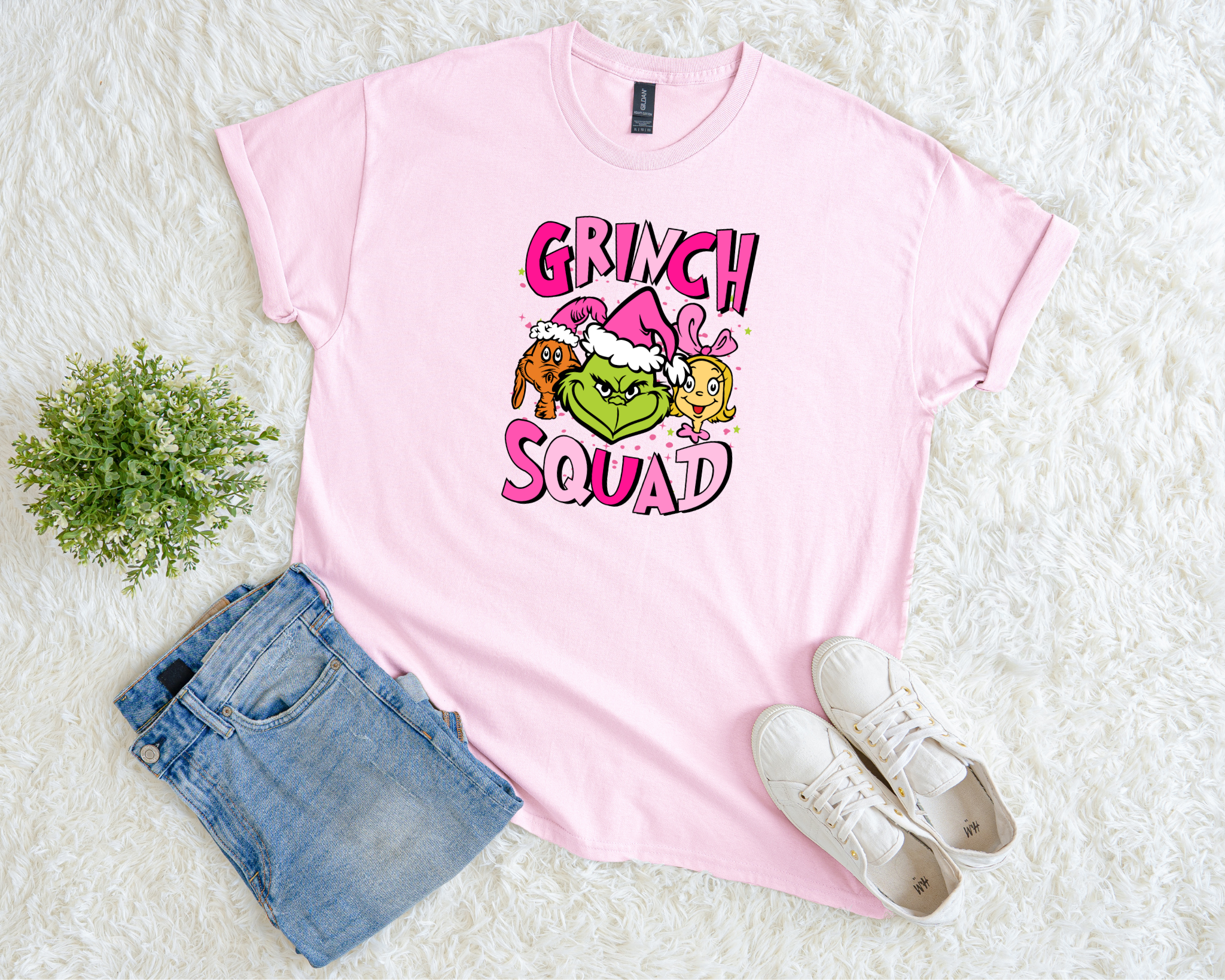 A light pink t-shirt featuring a colorful “Grinch Squad” Christmas design with the Grinch, Max, and Cindy Lou, styled flat with jeans and sneakers on a soft white background.