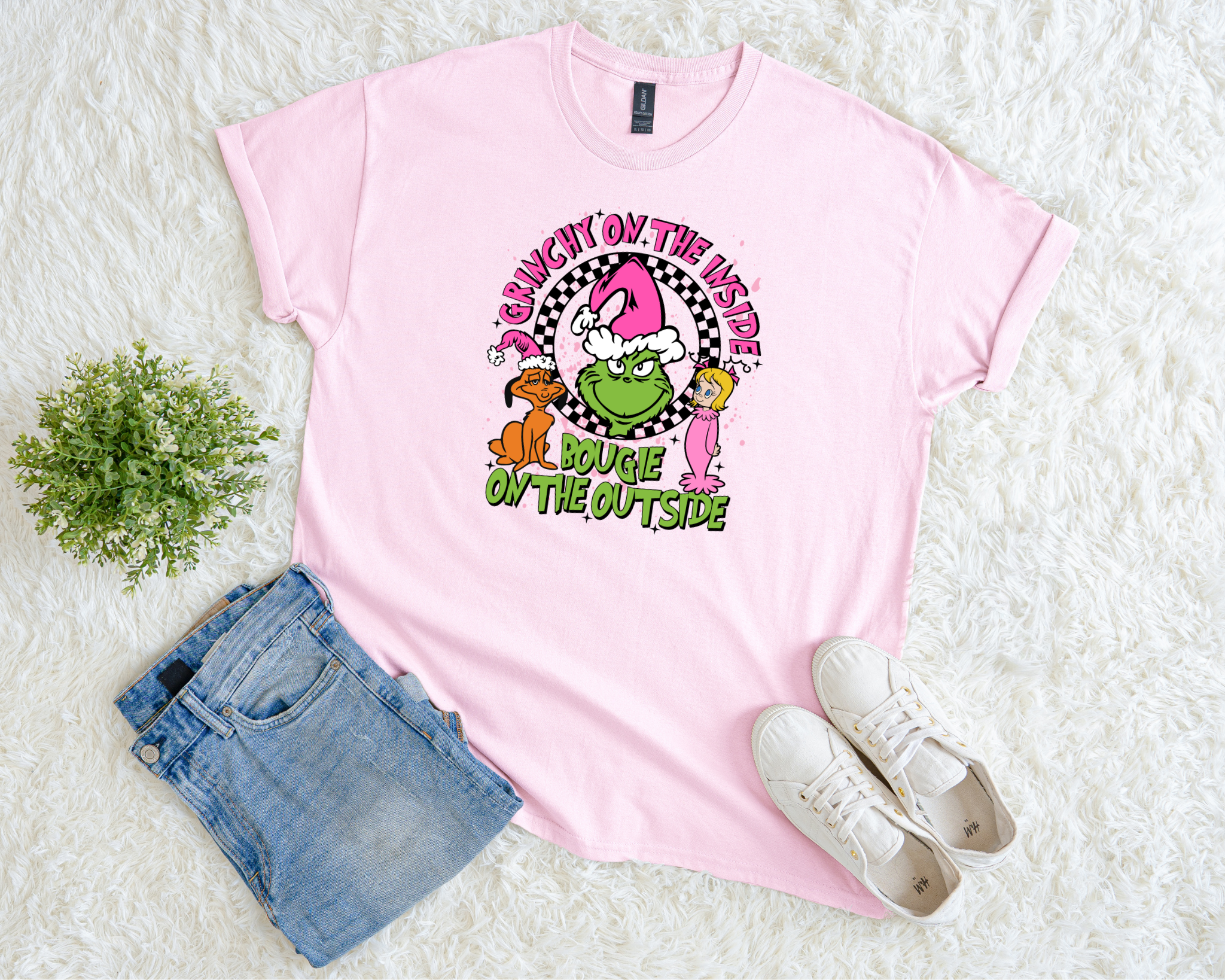 A light pink t-shirt featuring a playful “Grinchy on the Inside, Bougie on the Outside” Christmas design with pink, green, and black graphics, styled flat with jeans and sneakers on a white fluffy background.