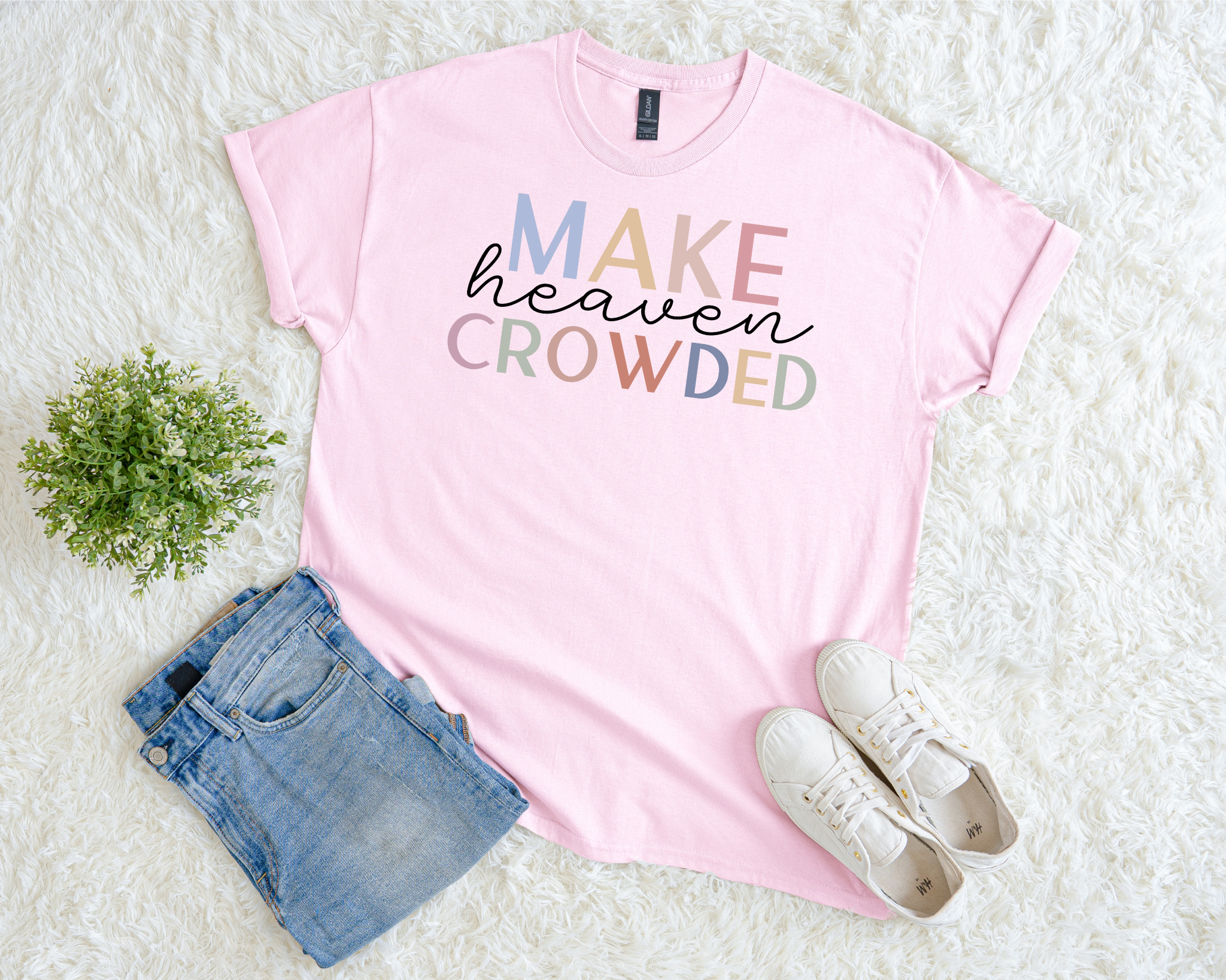 Pink t-shirt with 'MAKE heaven CROWDED' text, paired with denim shorts and white sneakers on a light background.