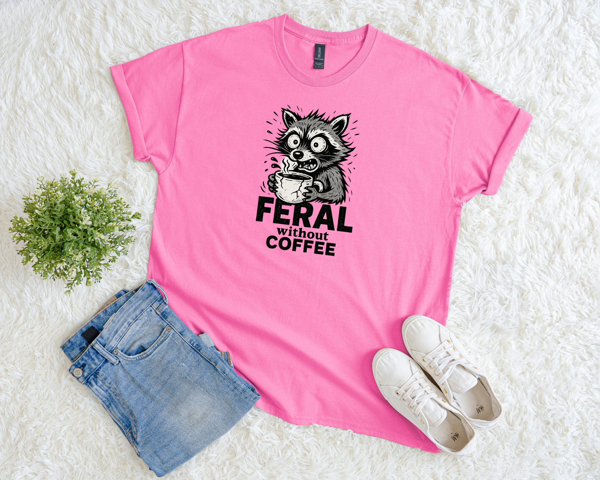 Pink t-shirt with a raccoon graphic feral without coffee, placed on a white surface with jeans and shoes