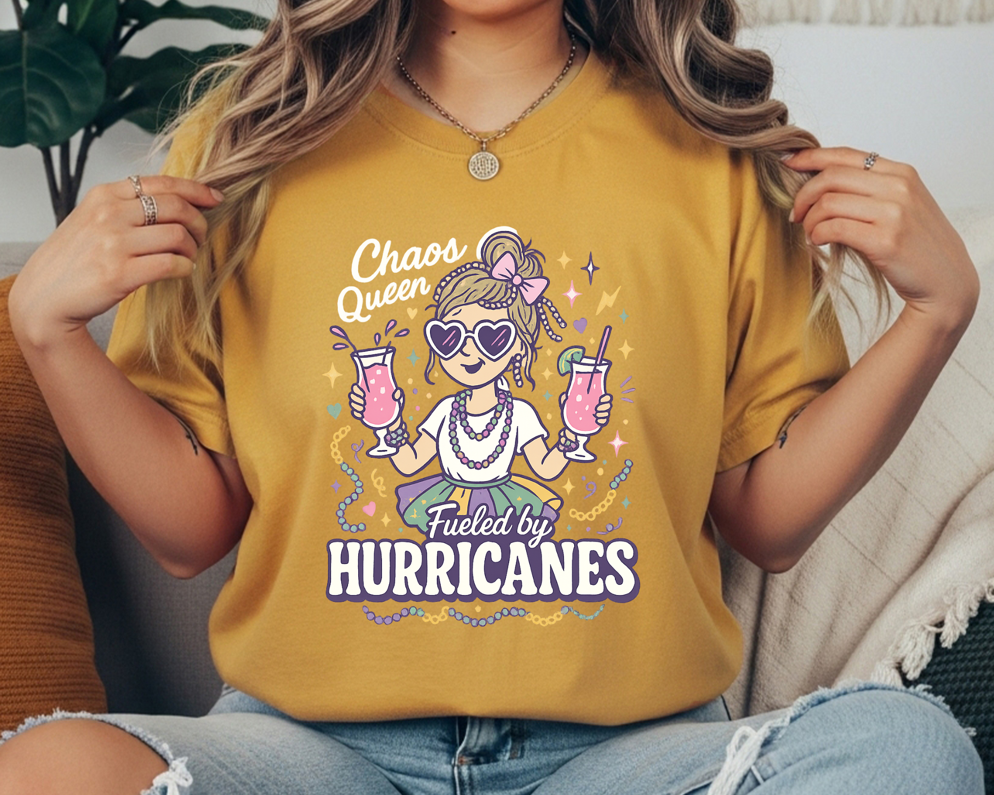mustard Comfort Colors Mardi Gras t-shirt with a playful girl holding two pink hurricane drinks, surrounded by beads and sparkles, and the text “Chaos Queen Fueled by Hurricanes