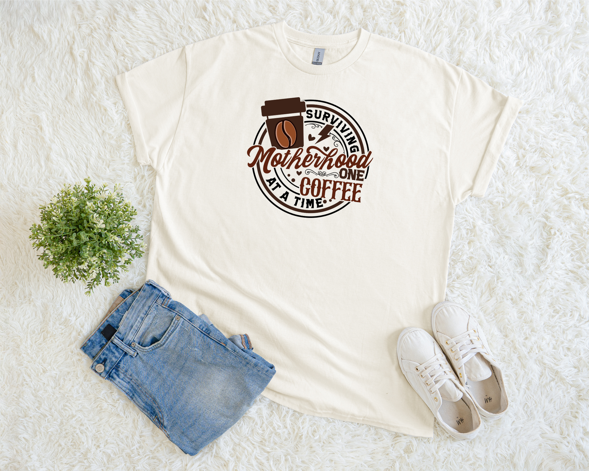 t-shirt featuring the phrase “Surviving Motherhood One Coffee at a Time” in brown lettering with a coffee cup graphic.