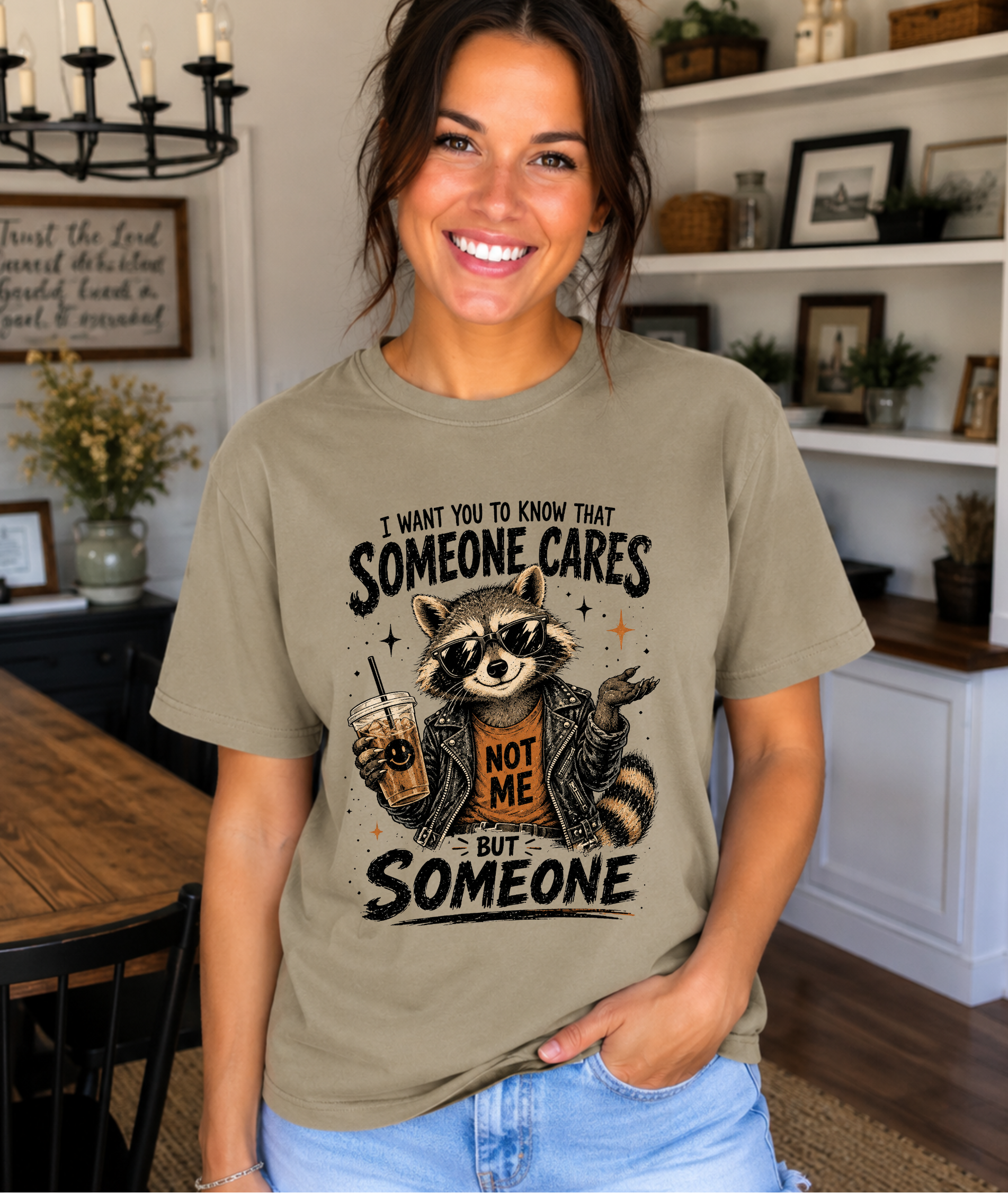 A woman wearing a graphic t-shirt in a warm, decorated indoor setting. The t-shirt is a muted olive-tan color with a black and brown graphic of a raccoon holding a drink, accompanied by the text 'I WANT YOU TO KNOW THAT SOMEONE CARES NOT ME BUT SOMEONE'.