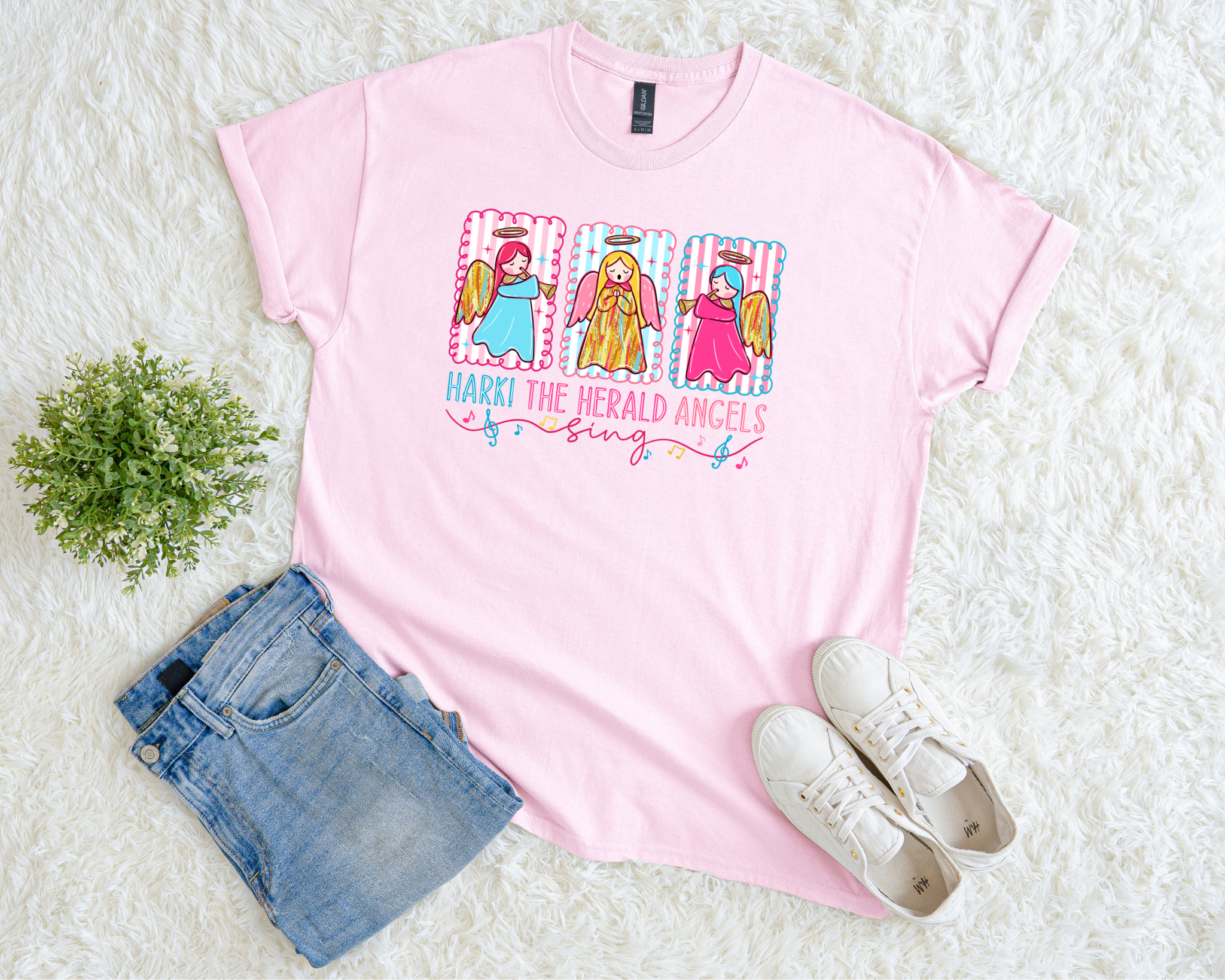 pink Christmas t-shirt featuring three colorful angels with gold wings and the phrase “Hark! The Herald Angels Sing,” styled flat with jeans and sneakers on a soft white background.