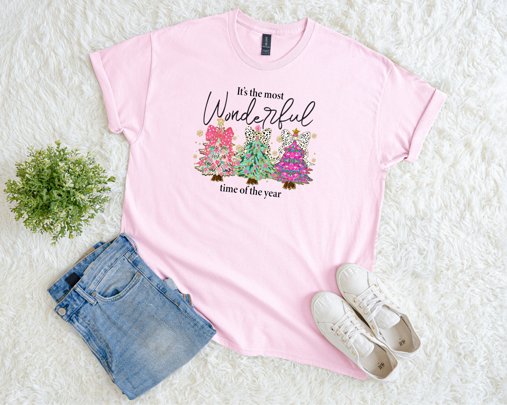pink Christmas t-shirt featuring three colorful trees with leopard bows and the phrase “It’s the most wonderful time of the year,” styled flat with jeans and sneakers on a soft white background.