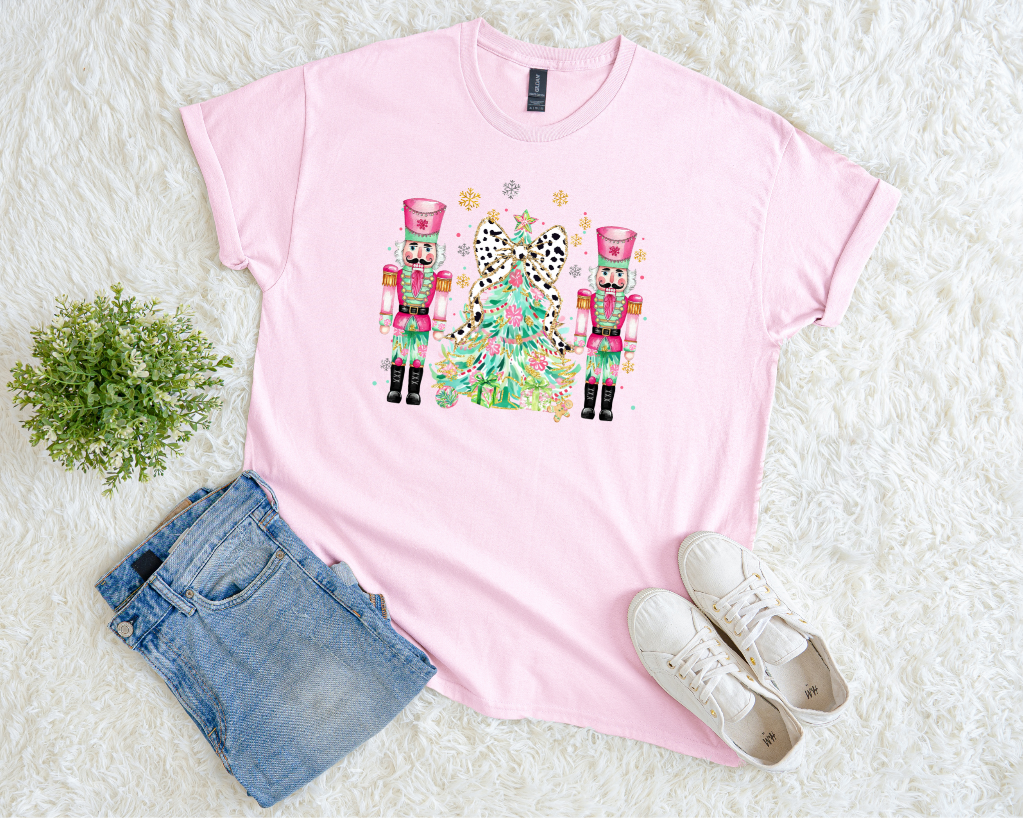 pink Christmas t-shirt featuring two pink nutcrackers and a leopard bow tree surrounded by snowflakes, styled flat with jeans and sneakers on a white fuzzy background.