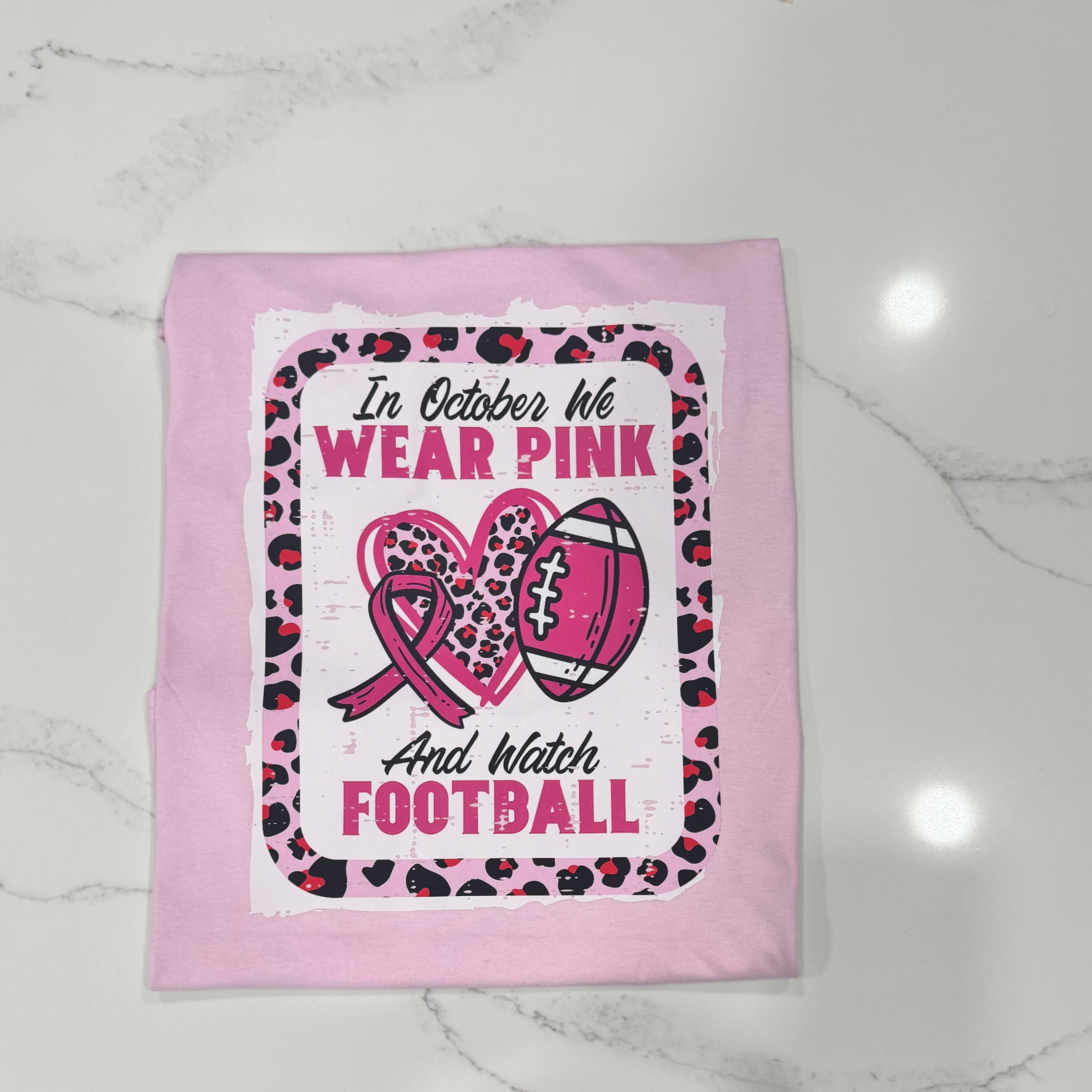 pink breast cancer awareness t-shirt with a leopard print border, football and pink ribbon design, and the phrase “In October We Wear Pink and Watch Football.”eRender