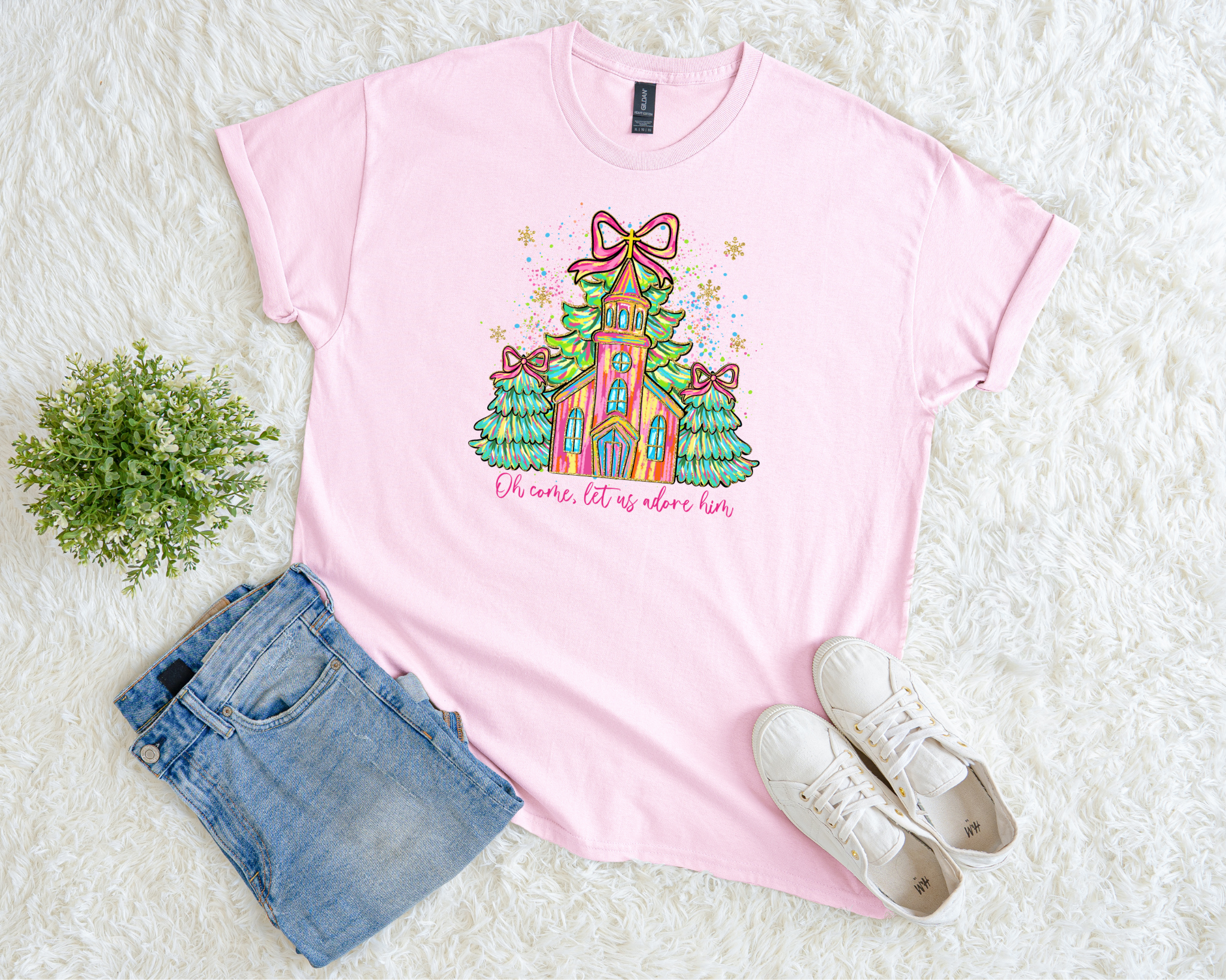 pink t-shirt featuring a colorful hand-drawn church surrounded by green Christmas trees with bows and the phrase “Oh Come, Let Us Adore Him,” styled flat with jeans and white sneakers.