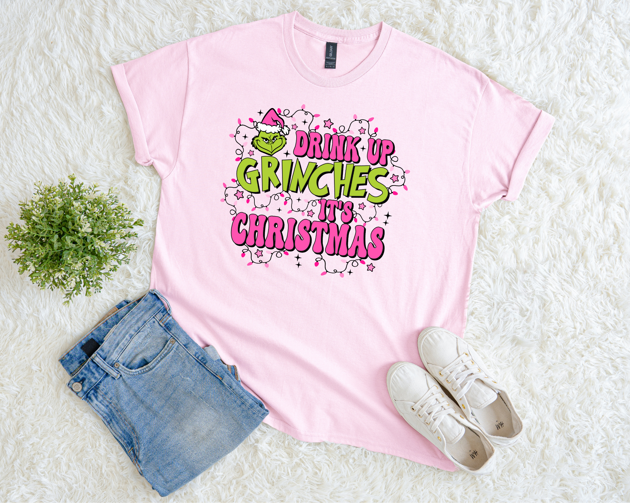 A light pink t-shirt featuring a playful “Drink Up Grinches, It’s Christmas” design in green and pink with Christmas lights, styled flat with jeans and sneakers on a soft