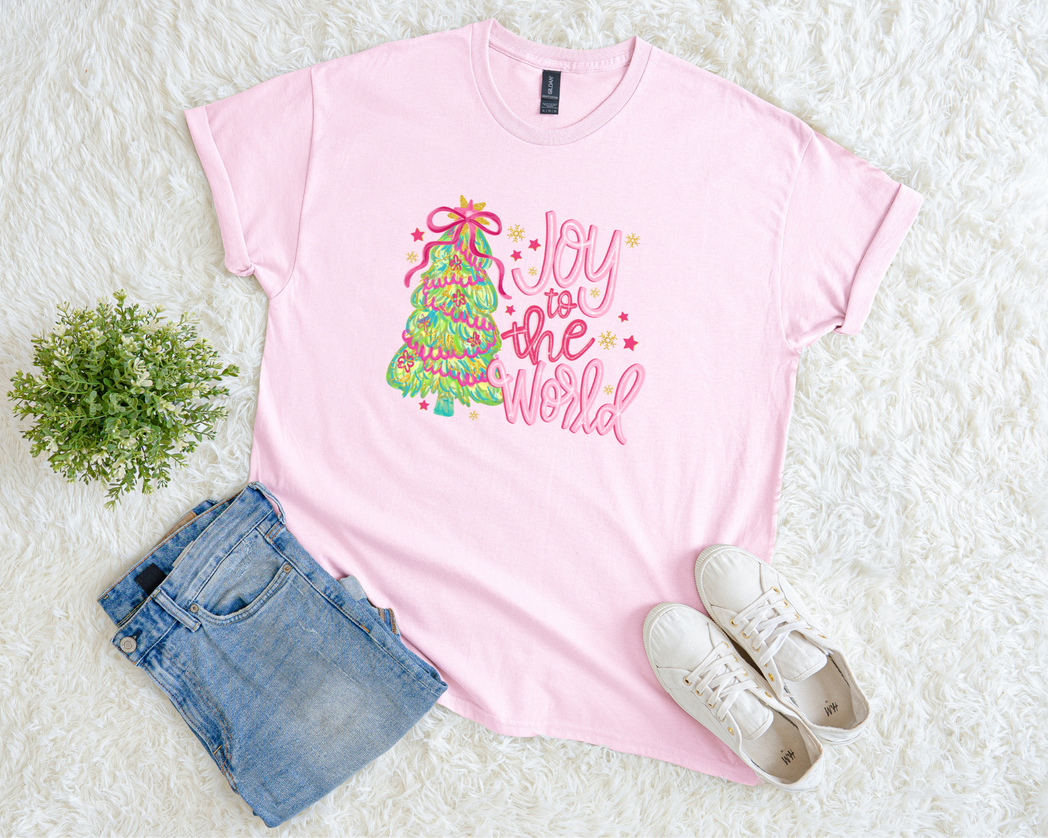 t-shirt with colorful “Joy to the World” design featuring a green and pink Christmas tree wrapped in ribbon, styled flat with jeans and white sneakers on a white textured background.