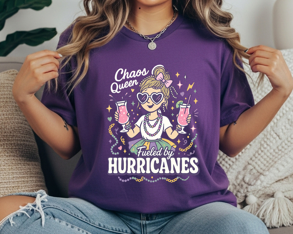 purple Comfort Colors Mardi Gras t-shirt with a playful girl holding two pink hurricane drinks, surrounded by beads and sparkles, and the text “Chaos Queen Fueled by Hurricanes