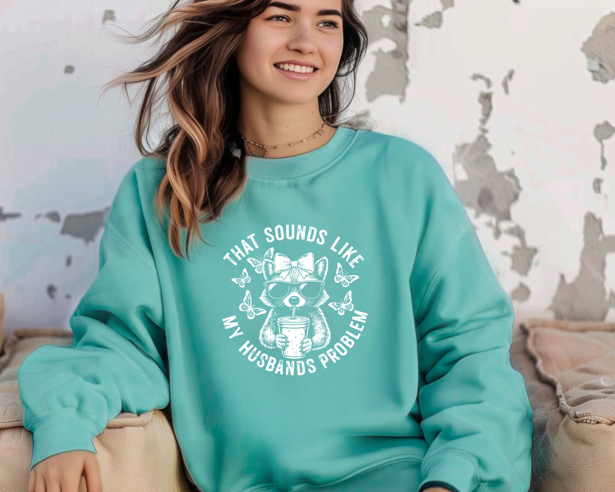crewneck sweatshirt with white circular graphic of a bow-wearing raccoon in sunglasses holding iced coffee, surrounded by butterflies, reading “That Sounds Like My Husband’s Problem.”