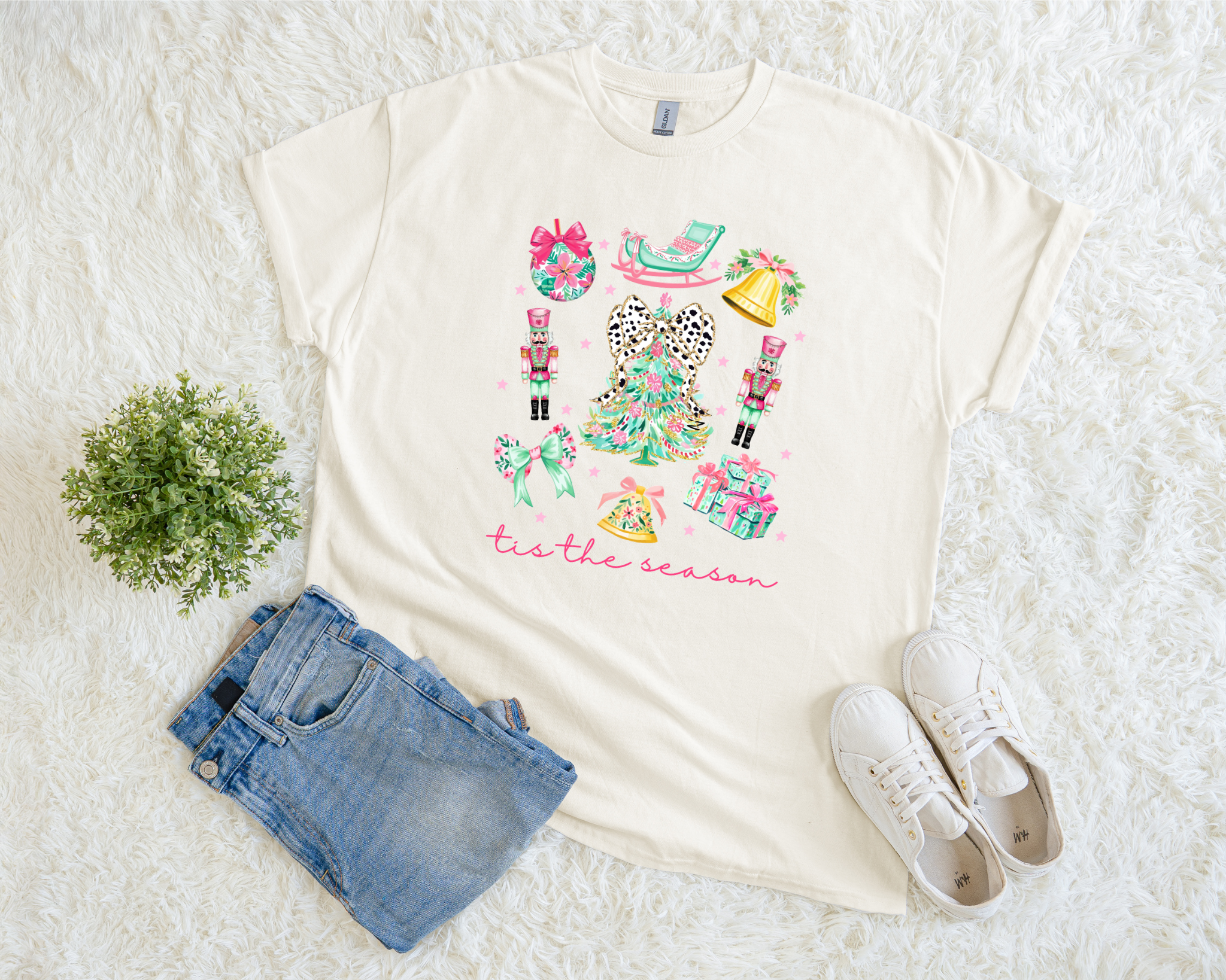 t-shirt featuring a colorful “‘Tis the Season” design with nutcrackers, pastel gifts, bows, and a leopard-bow Christmas tree, styled flat with jeans and white sneakers.