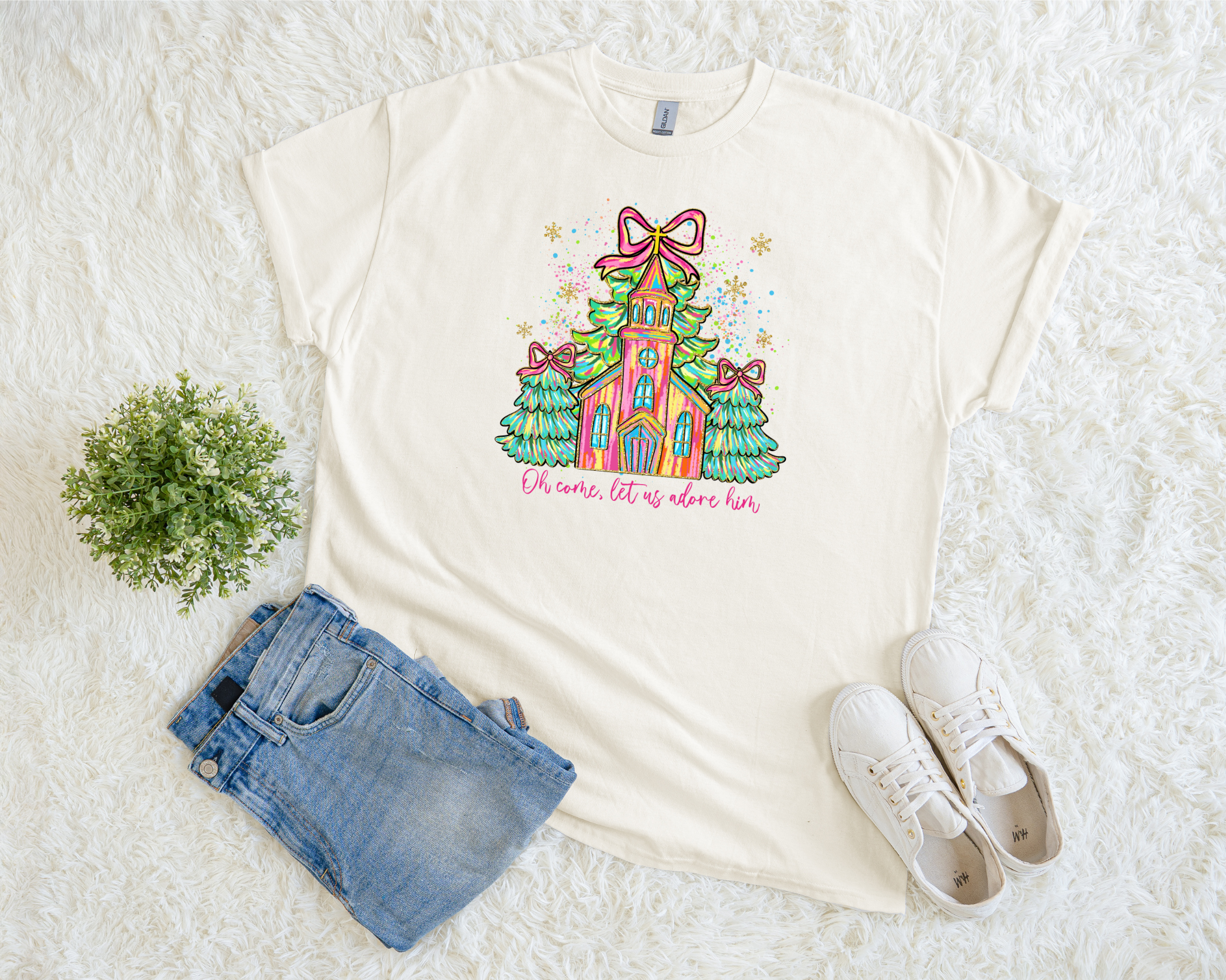 Cream-colored t-shirt featuring a colorful hand-drawn church surrounded by green Christmas trees with bows and the phrase “Oh Come, Let Us Adore Him,” styled flat with jeans and white sneakers.
