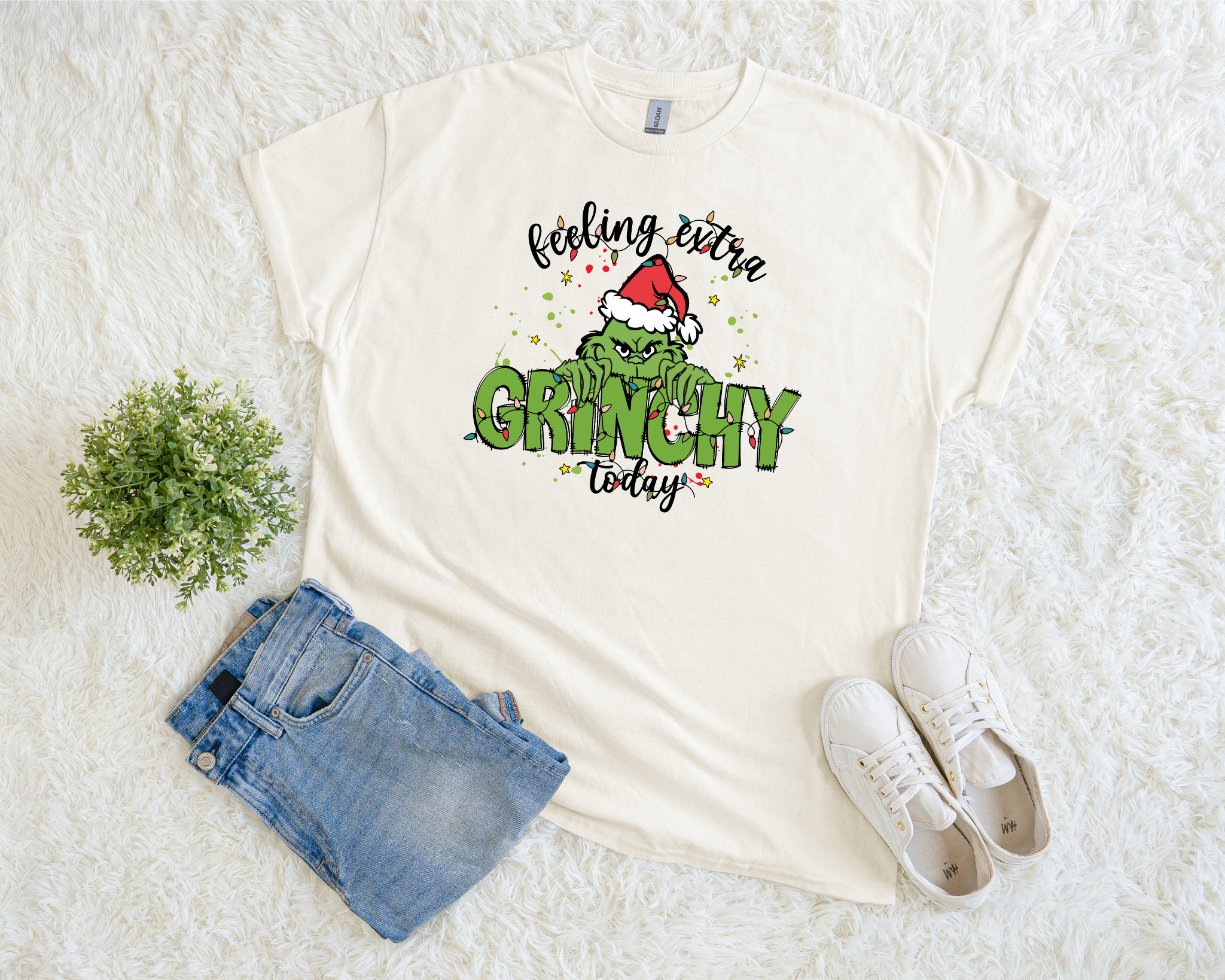 A cream-colored t-shirt featuring a festive “Feeling Extra Grinchy Today” Christmas design with green lettering and a Santa hat graphic, styled flat with jeans and sneakers on a soft white background.