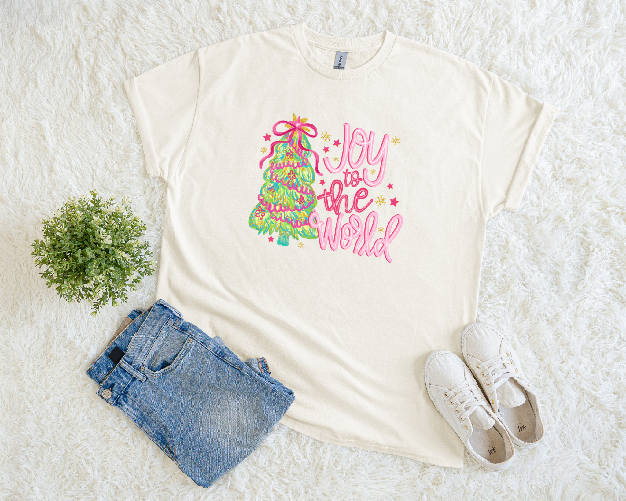 t-shirt with colorful “Joy to the World” design featuring a green and pink Christmas tree wrapped in ribbon, styled flat with jeans and white sneakers on a white textured background.