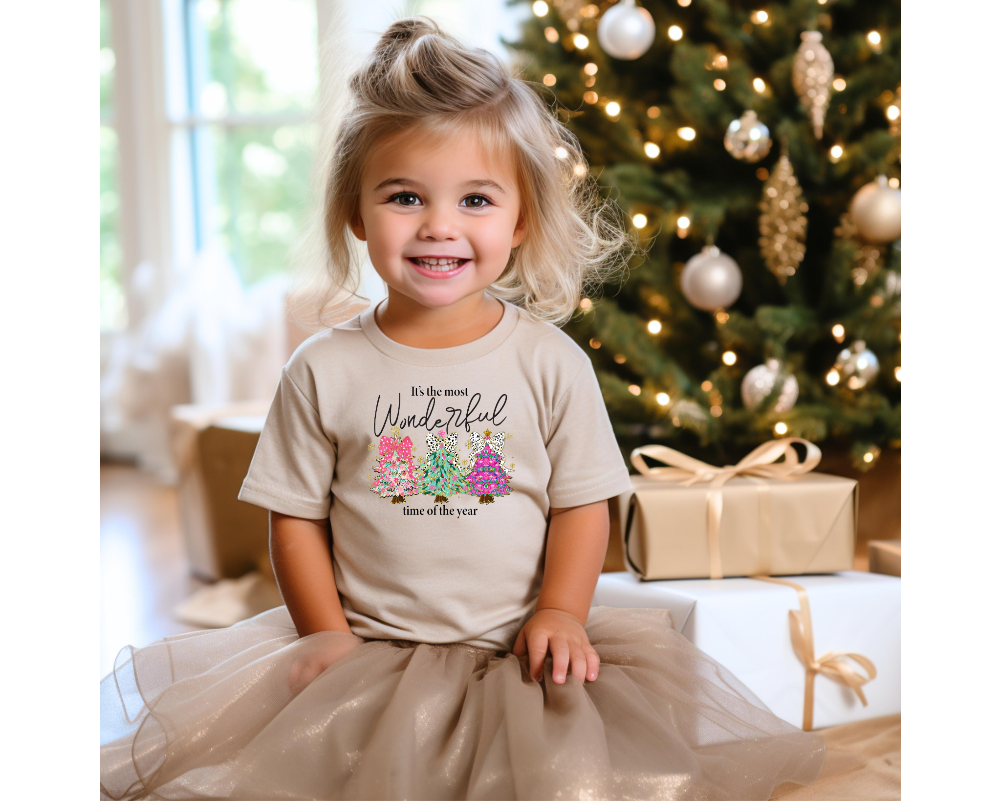 A young girl wearing a tan Christmas t-shirt with colorful trees and leopard bows that reads “It’s the Most Wonderful Time of the Year,” sitting happily near a decorated Christmas tree with presents.