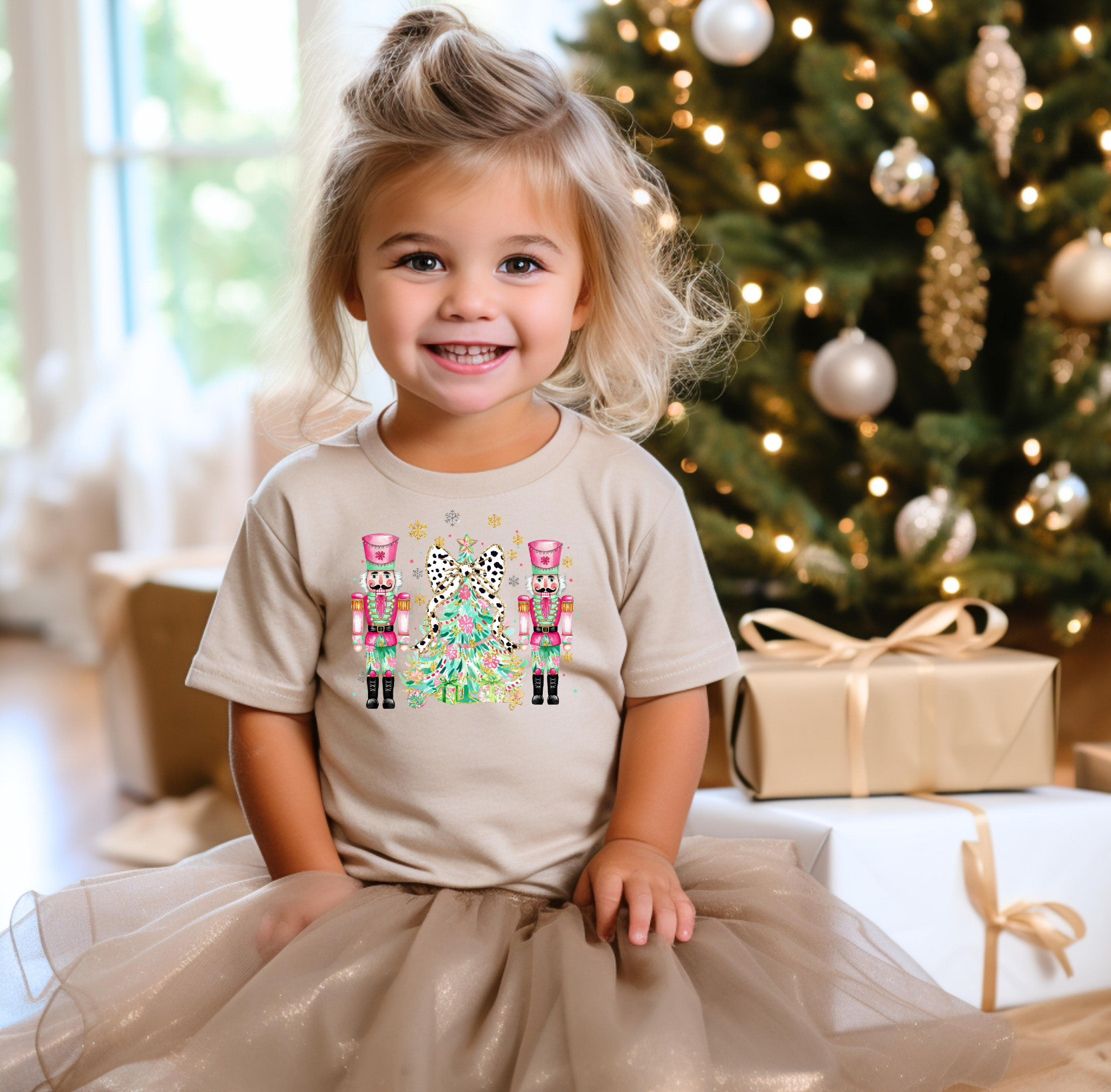 A young girl wearing a tan Nutcracker Christmas t-shirt featuring two pink nutcrackers and a leopard bow Christmas tree, smiling while sitting near a decorated Christmas tree with wrapped presents.