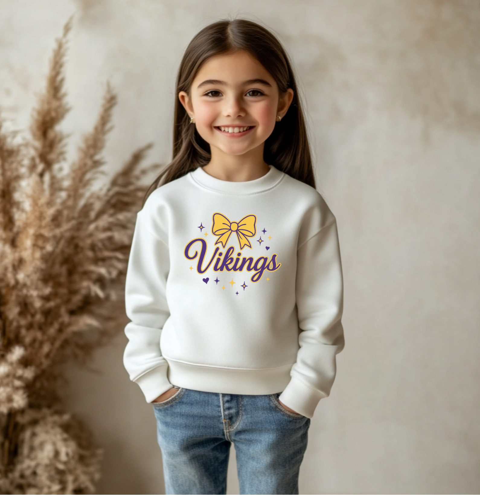 Young girl wearing a white sweatshirt with 'Vikings' and a bow design, standing against a neutral background.