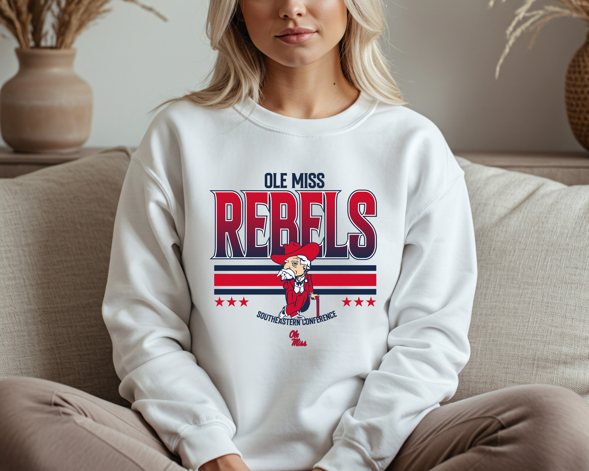 A woman wearing a light gray Ole Miss Rebels-inspired crewneck sweatshirt with bold red and navy graphics and a vintage-style mascot design, sitting comfortably on a couch.